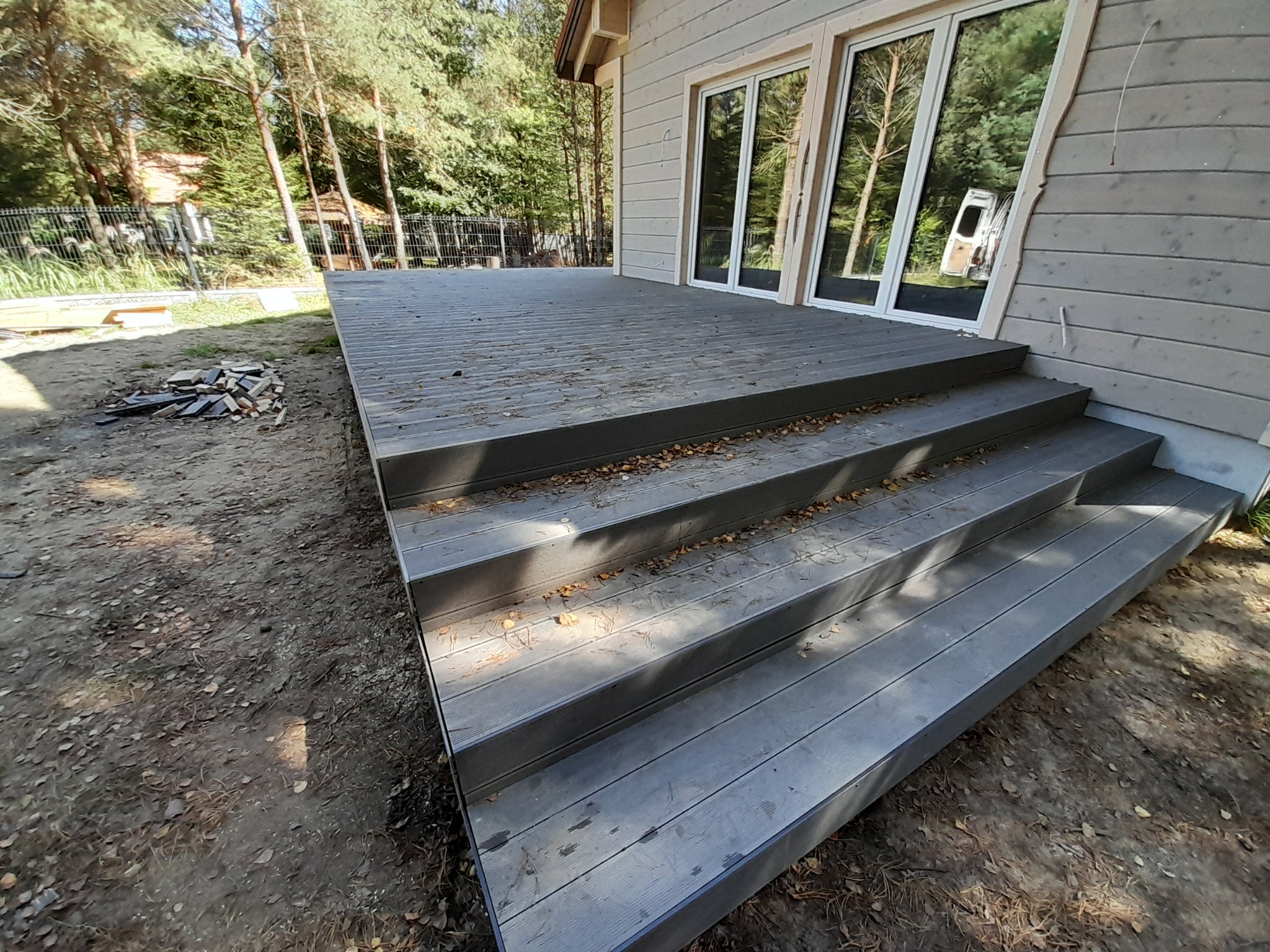 Nowoczesny taras kompozytowy z schodami, przylegający do domu z dużymi oknami, otoczony zielenią. Widoczne ślady liści na powierzchni tarasu.