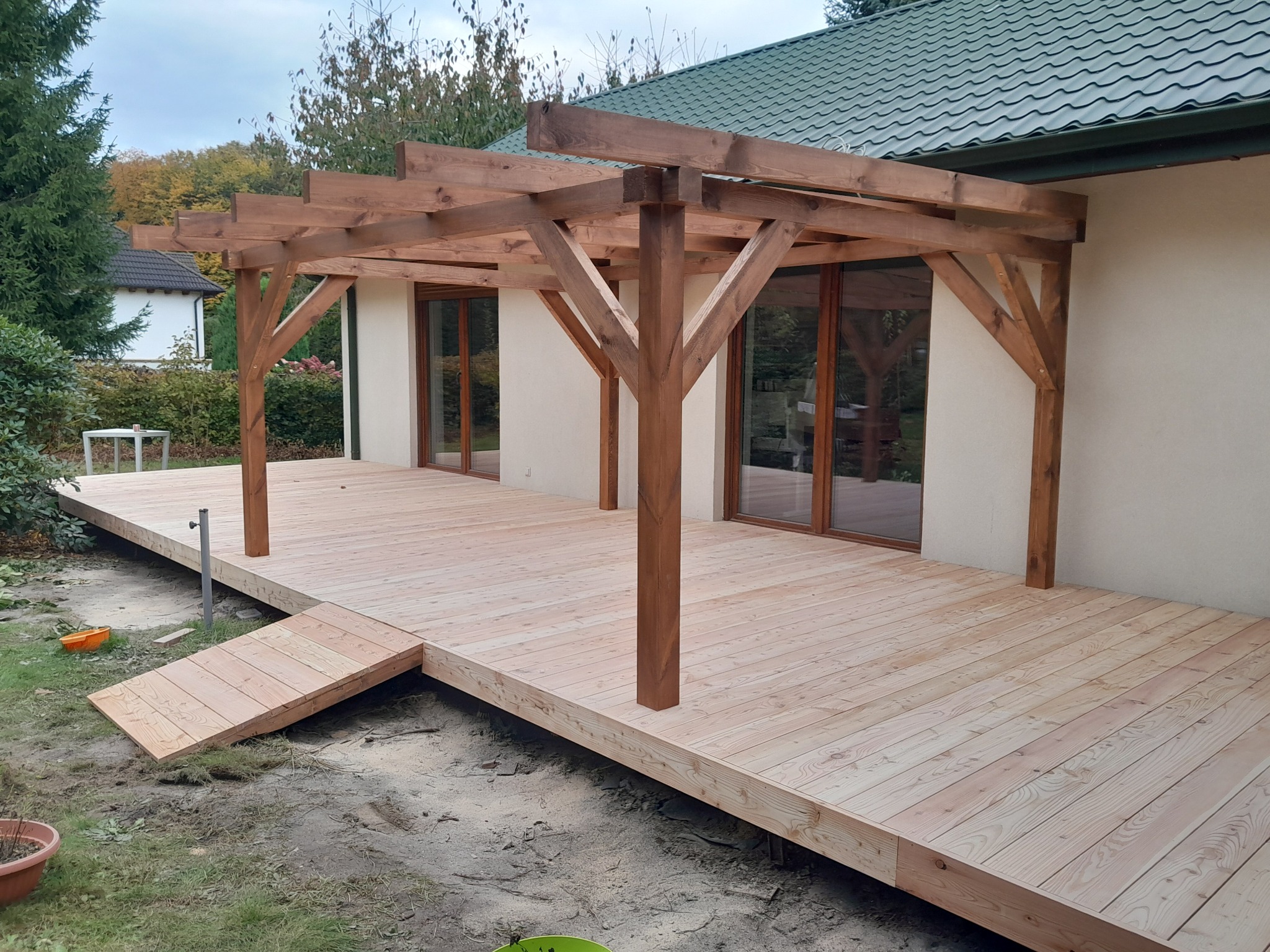 Jasny taras drewniany z pergolą, widok z poziomu gruntu. Drewniana rampa prowadzi na taras. W tle dom z zielonym dachem i drzewa.