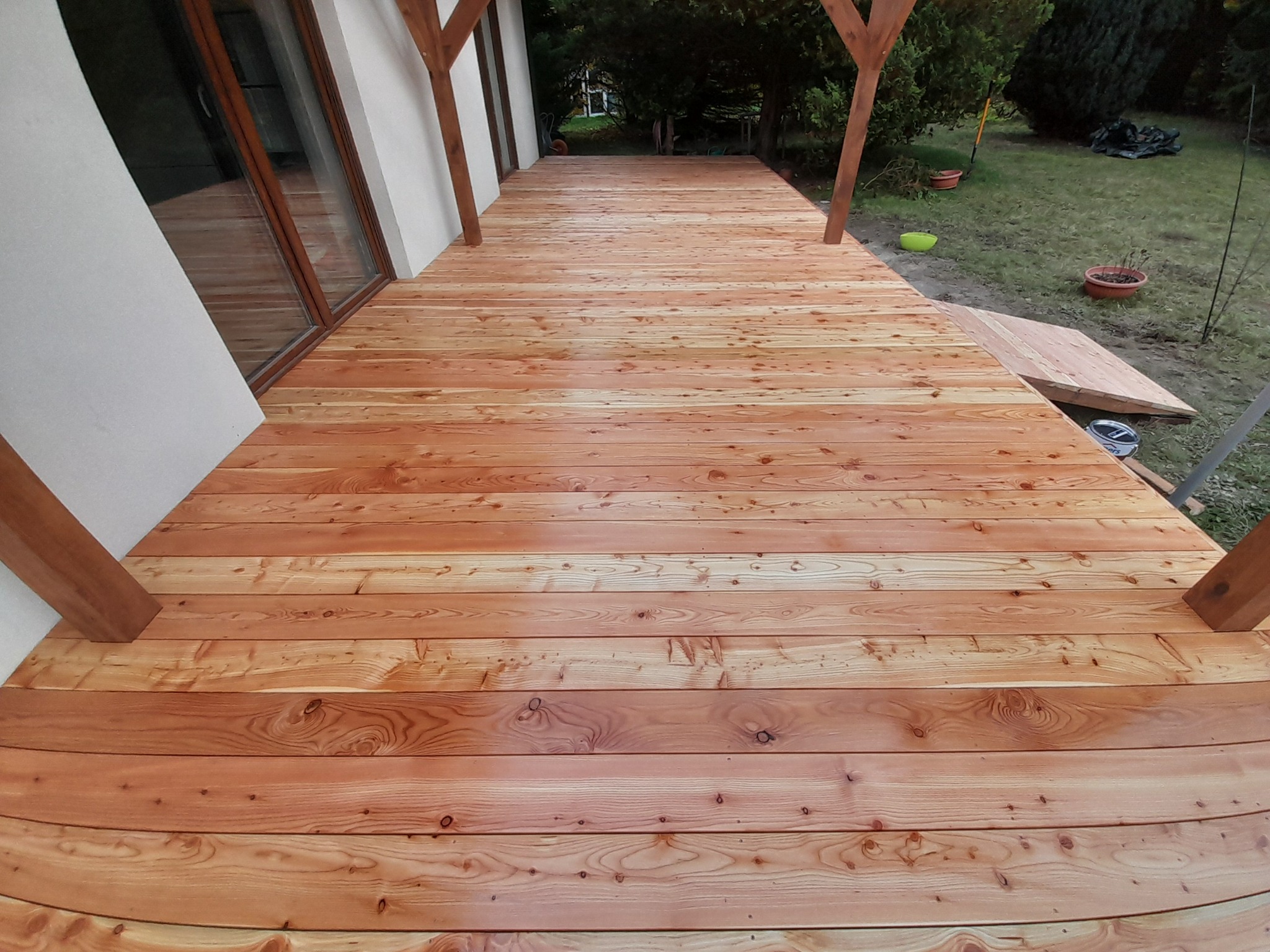 Jasny, drewniany taras z widoczną strukturą drewna, częściowo zadaszony, z elementami konstrukcyjnymi z drewna. Widok na ogród w tle.