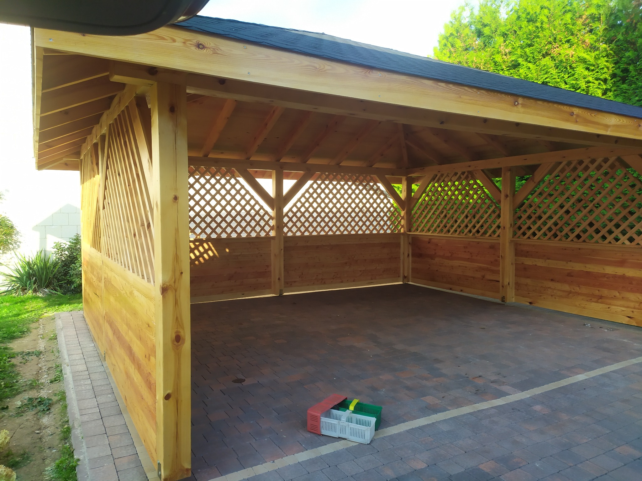 Drewniana altana z kratownicowymi ścianami, pokryta czarnym dachem, stoi na brukowanym podłożu. Na podłodze leżą kolorowe skrzynki.