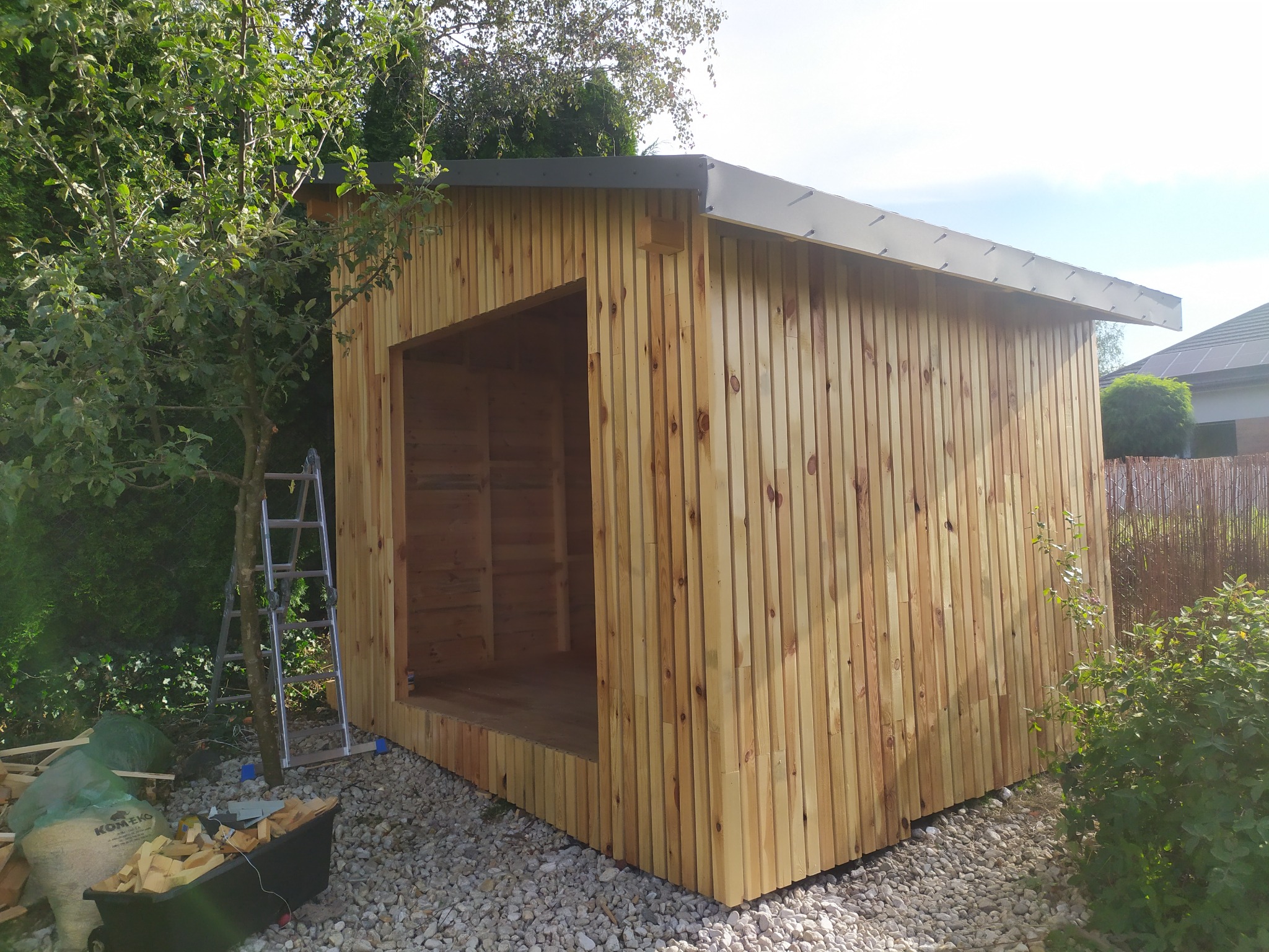 Surowa drewniana altana z szarą blachą na dachu, w trakcie budowy, z widoczną drabiną i workiem z drewnem na żwirowym podłożu, w otoczeniu zieleni.