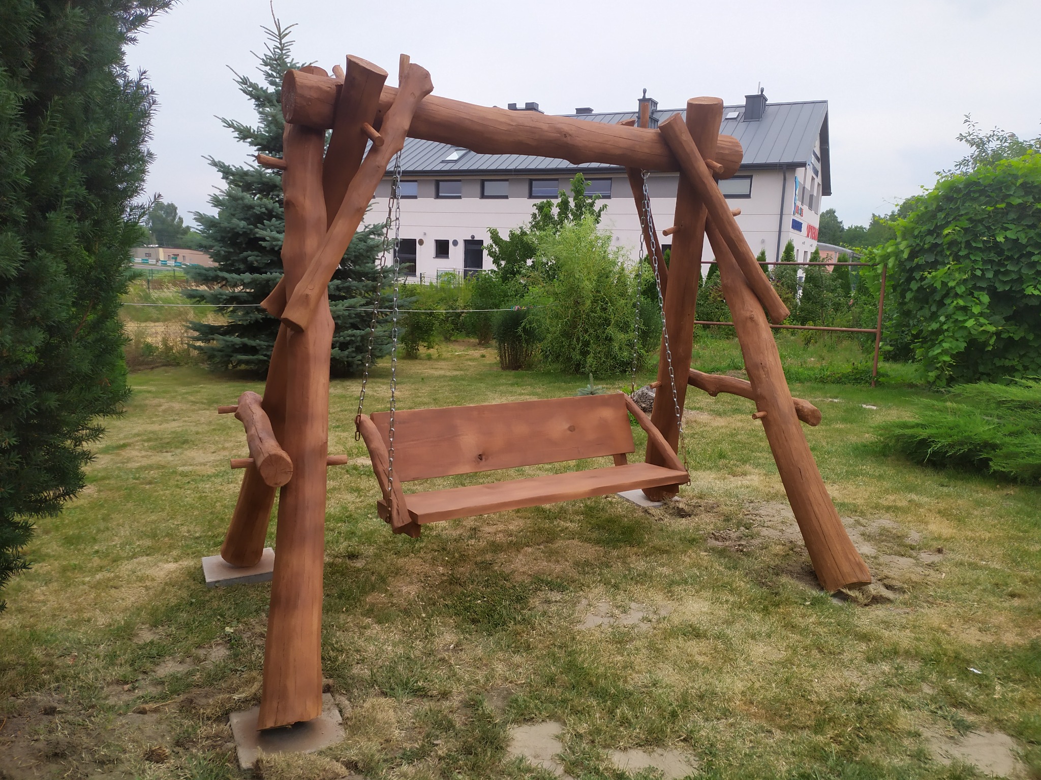 Drewniana huśtawka ogrodowa z bali, zawieszona na łańcuchach, na tle budynku i zieleni. Konstrukcja z surowego drewna, wkomponowana w naturalny krajobraz.