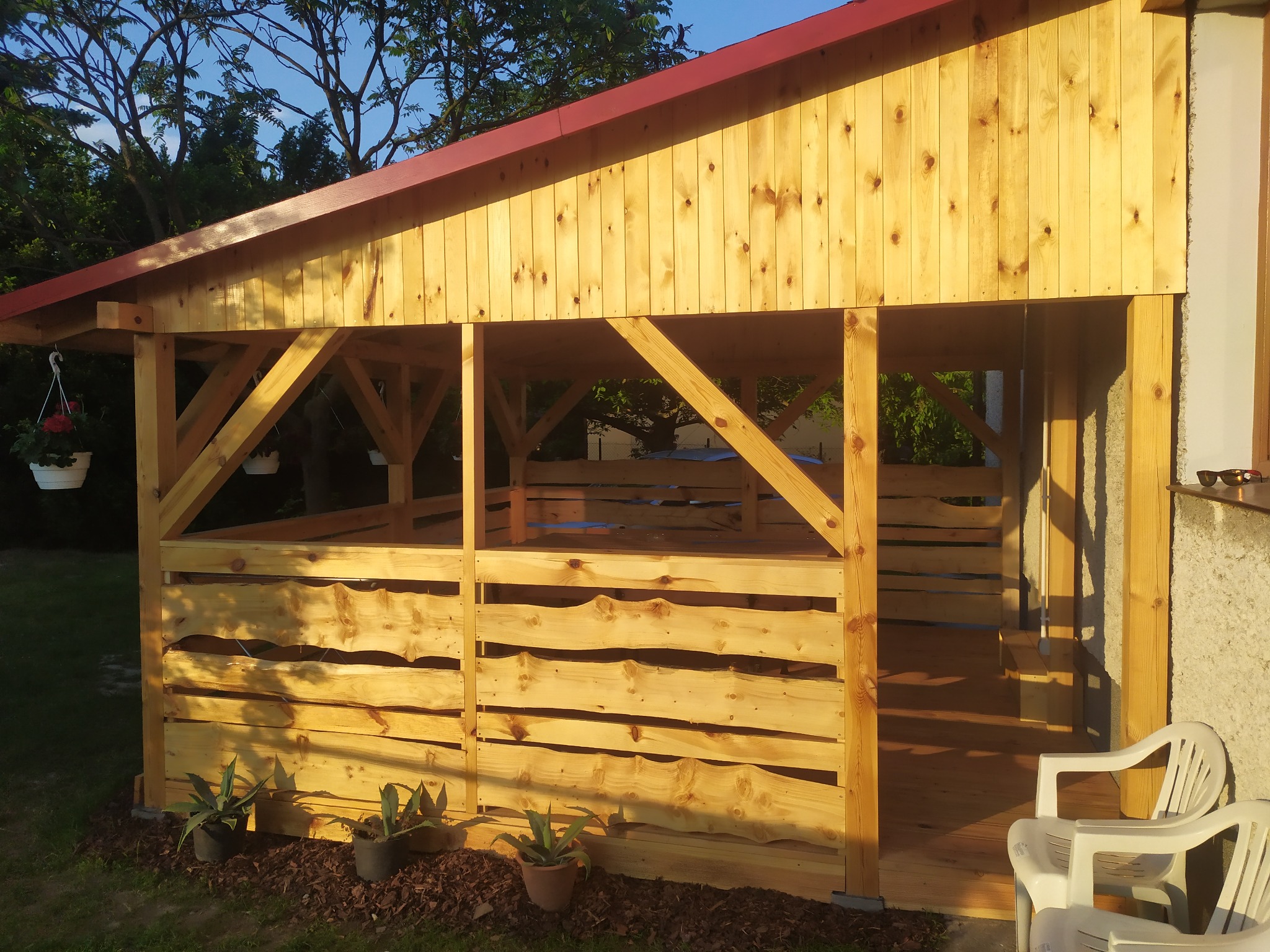 Drewniana altana z czerwonym dachem, wykonana z surowego drewna, z roślinami w doniczkach u podstawy i białymi krzesłami obok, w słoneczny dzień.