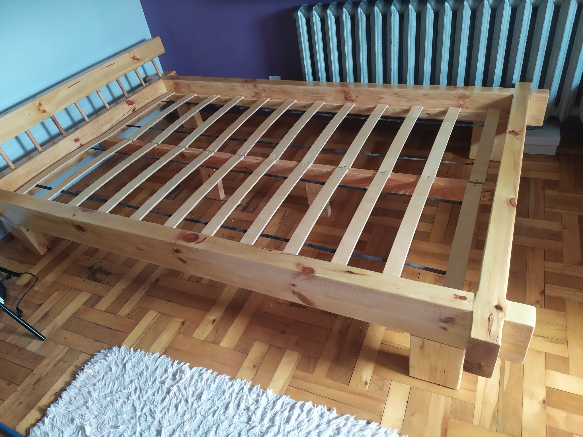 Surowa rama łóżka z jasnego drewna z listwami, widoczna na tle parkietu i grzejnika. Elementy łóżka są świeżo obrobione, z widocznymi słojami drewna.
