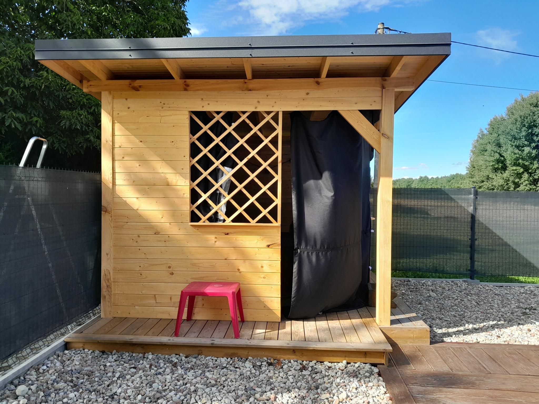 Drewniana altana z kratką w oknie i zasłoną, na podłożu z kamieni i drewnianym podeście. Czerwony stołek przed wejściem. Widok na ogród w tle.