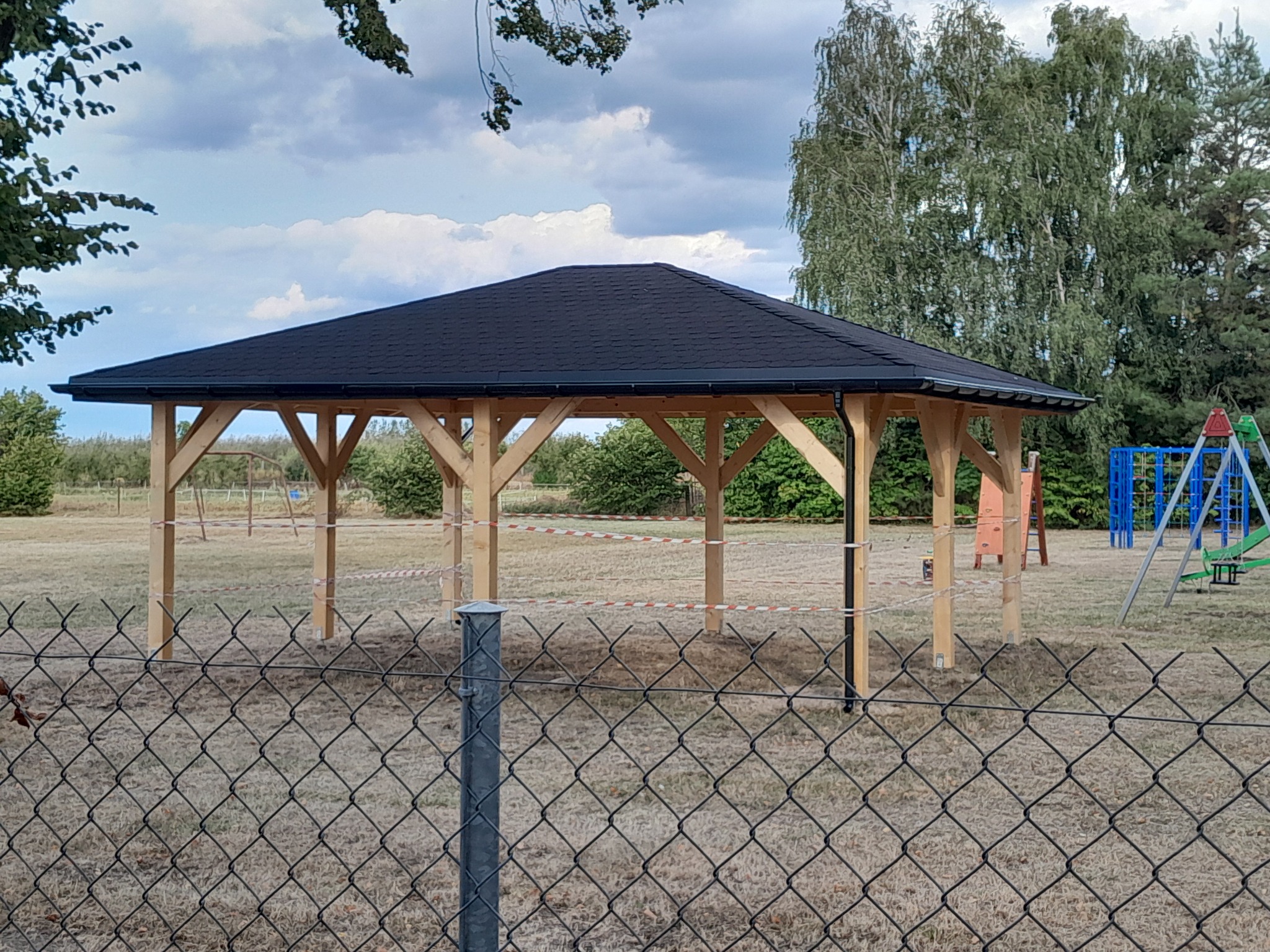 Nowa, drewniana altana z czarnym dachem, widoczna przez siatkę ogrodzeniową. W tle plac zabaw i drzewa, pochmurne niebo.