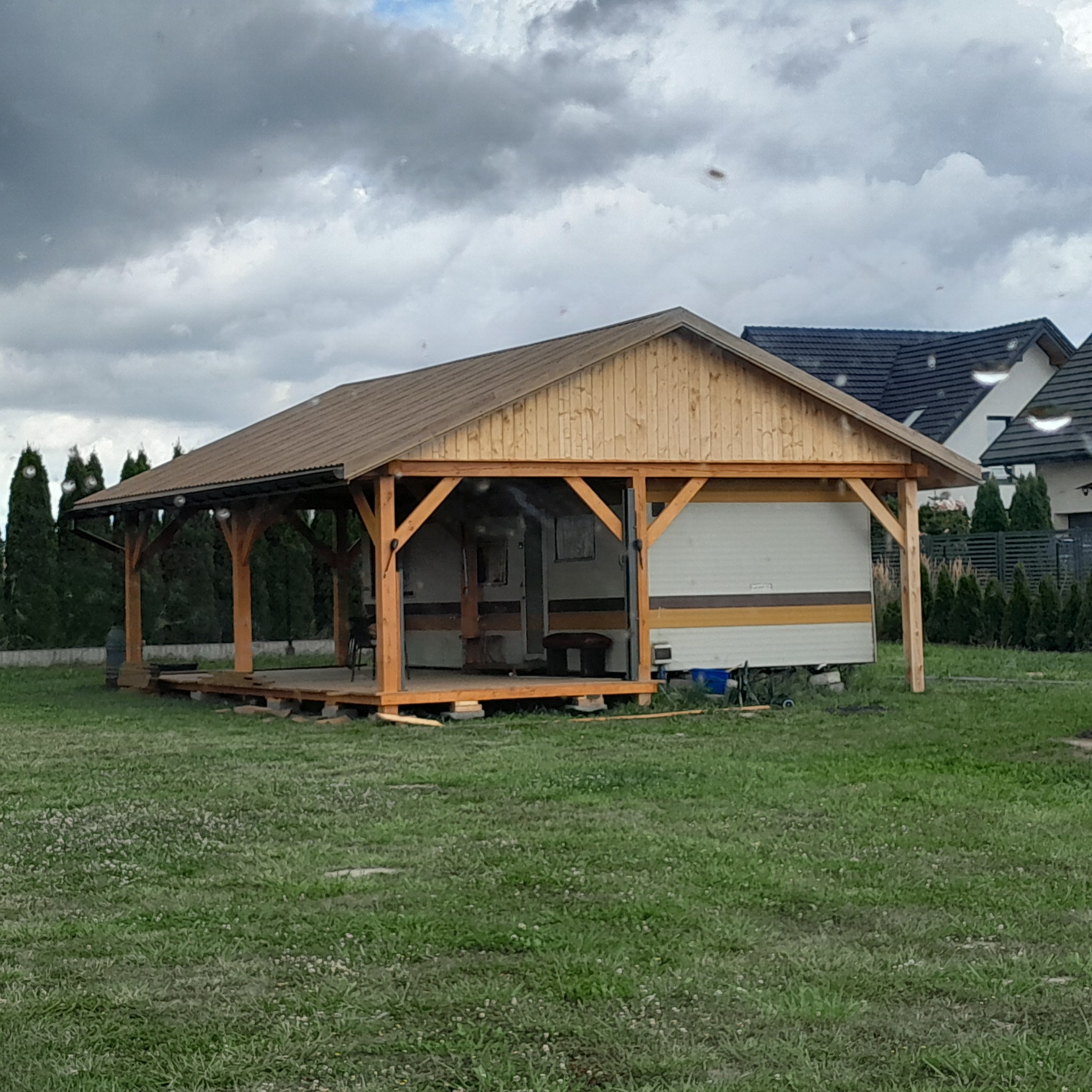 Drewniana altana z brązowym dachem, częściowo osłaniająca przyczepę kempingową na zielonym trawniku w pochmurny dzień. Widoczne elementy konstrukcyjne i wykończeniowe.