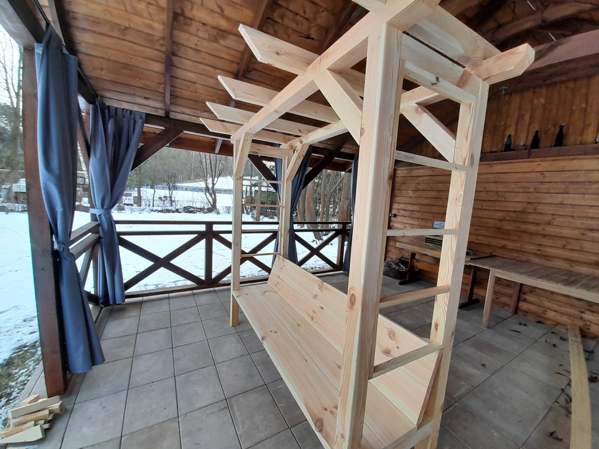 Drewniana pergola z ławką w trakcie montażu na zadaszonym tarasie z szarymi zasłonami. Widoczne elementy konstrukcyjne i fragment zimowego krajobrazu w tle.