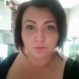 Portret kobiety z ciemnymi włosami, z widocznym kolczykiem w brwi i wardze, stojącej w pomieszczeniu z rozmytym tłem, na którym widać szafki i rośliny.