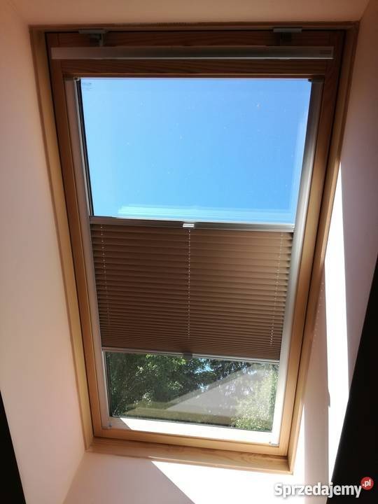 Beżowe plisy okienne częściowo zasłonięte w oknie dachowym, widok na błękitne niebo i zieleń drzew z Nowego Sącza.