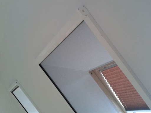 Zasłonięte okno dachowe z plisowaną roletą w drewnianej ramie, widok z dołu, białe ściany.