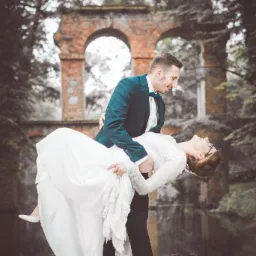 Pan młody trzyma pannę młodą w ramionach na tle ceglanego łuku i drzew, ona w białej sukni z koronkowymi rękawami i szpilkach, on w ciemnym garniturze z muszką.