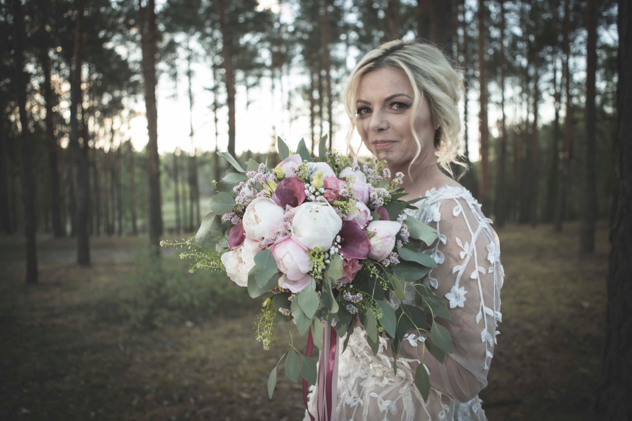 Panna młoda w koronkowej sukni trzyma bukiet z białych i różowych piwonii oraz eukaliptusa na tle leśnej scenerii.