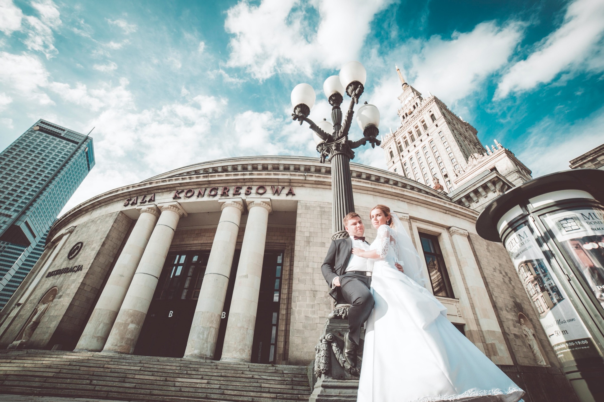 Para młoda pozuje na tle Sali Kongresowej w Warszawie, on w czarnym garniturze z muszką, ona w białej sukni ślubnej z koronkowymi rękawami, szeroki kadr z perspektywy żabiej, widok na budynki...