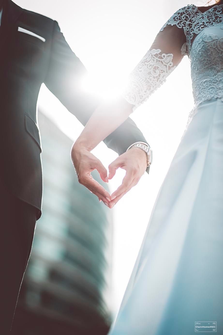 Nowożeńcy trzymają się za ręce, formując kształt serca na tle rozświetlonego wieżowca w słoneczny dzień. Panna młoda w koronkowej sukni z długim rękawem, pan młody w czarnym garniturze i srebrnym...