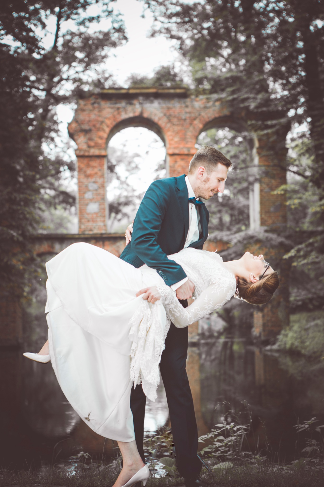 Pan młody trzyma pannę młodą w ramionach na tle ceglanego łuku i drzew, ona w białej sukni z koronkowymi rękawami i szpilkach, on w ciemnym garniturze z muszką.