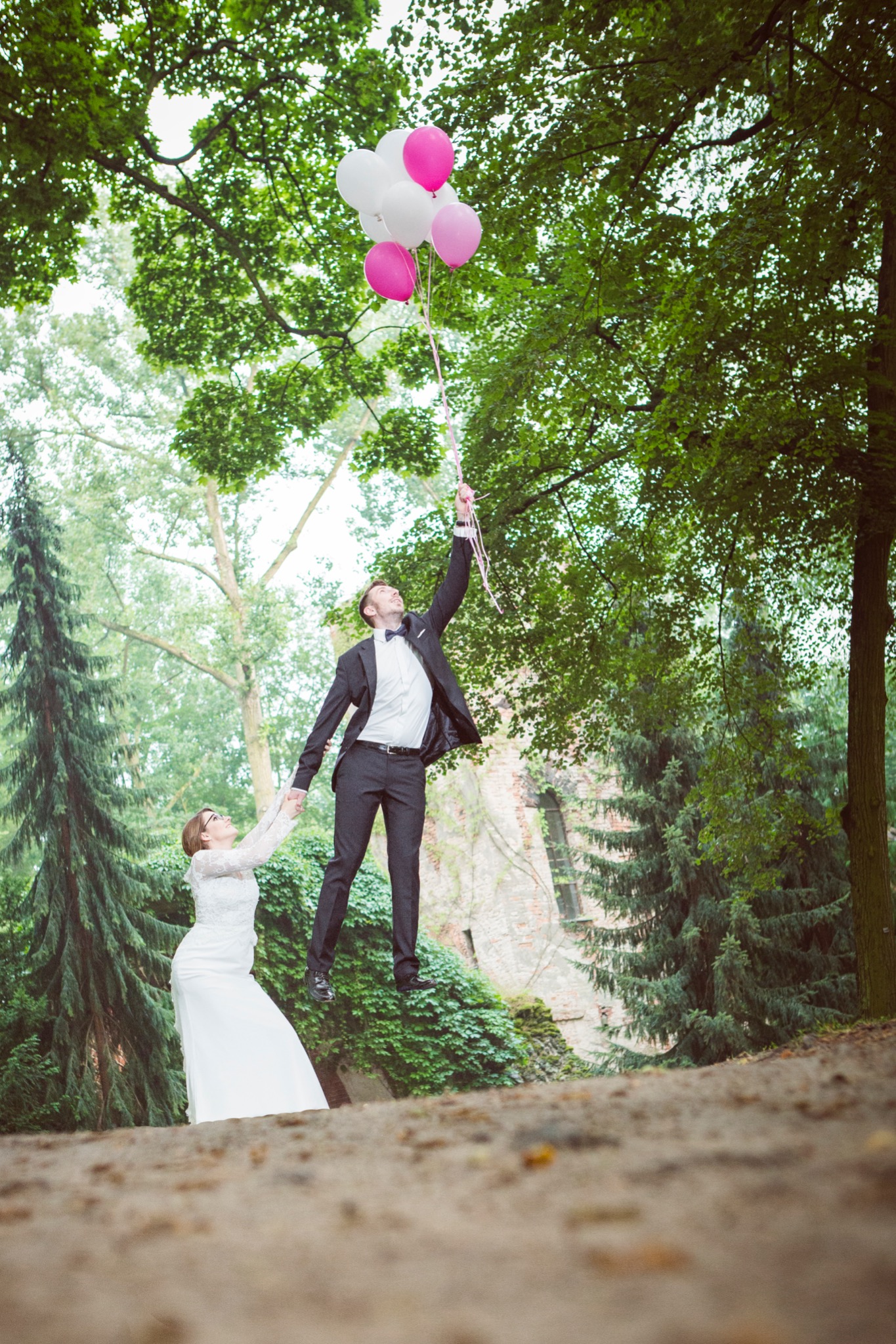 Para młoda na zewnątrz: panna młoda w białej sukni trzyma za rękę pana młodego w garniturze, który unosi się w powietrzu trzymając pęk białych i różowych balonów na tle zieleni drzew i muru...