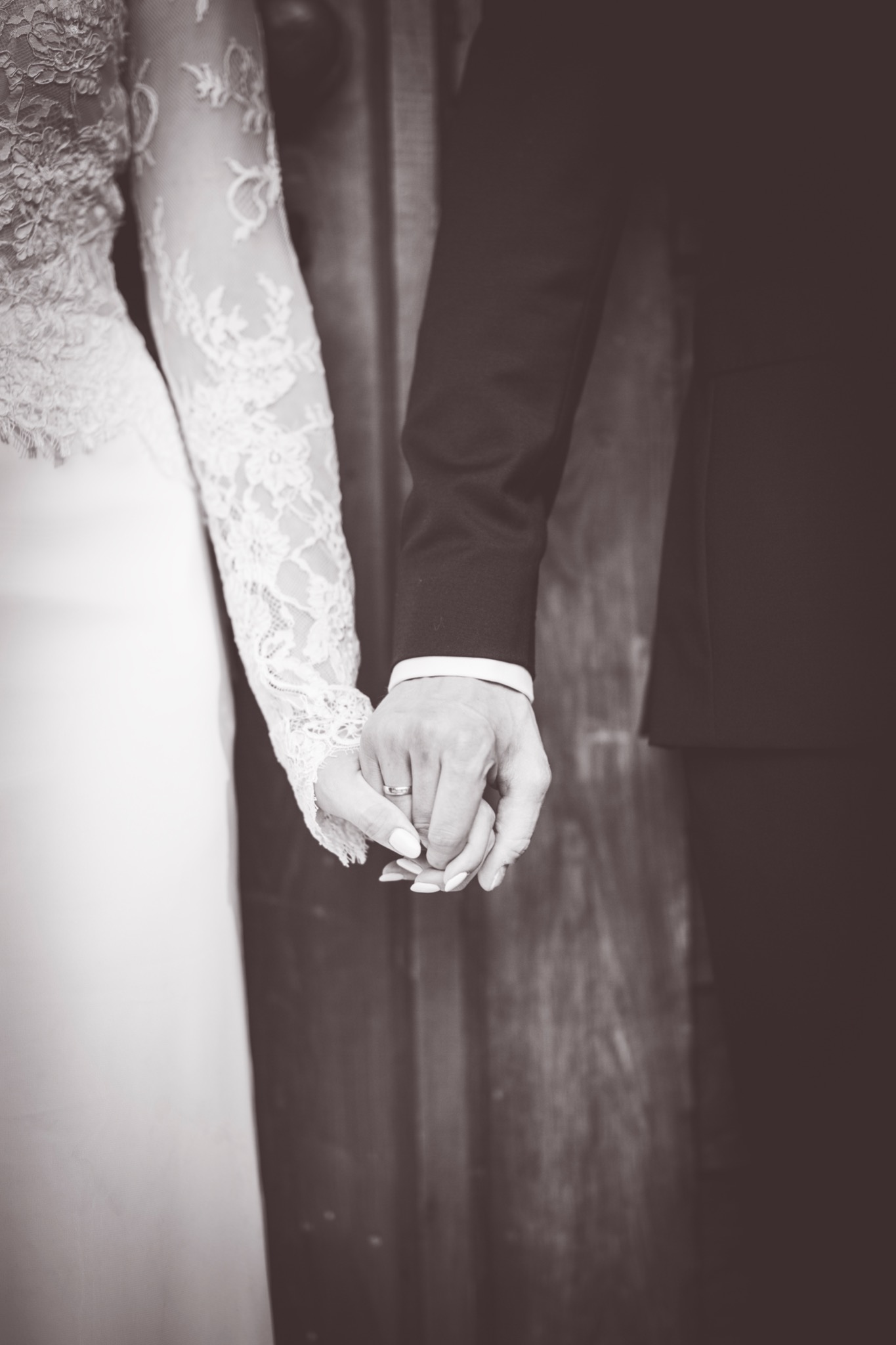 Czarno-białe zbliżenie dłoni pary młodej trzymających się za ręce, widoczny pierścionek na palcu panny młodej i koronkowy rękaw sukni ślubnej.