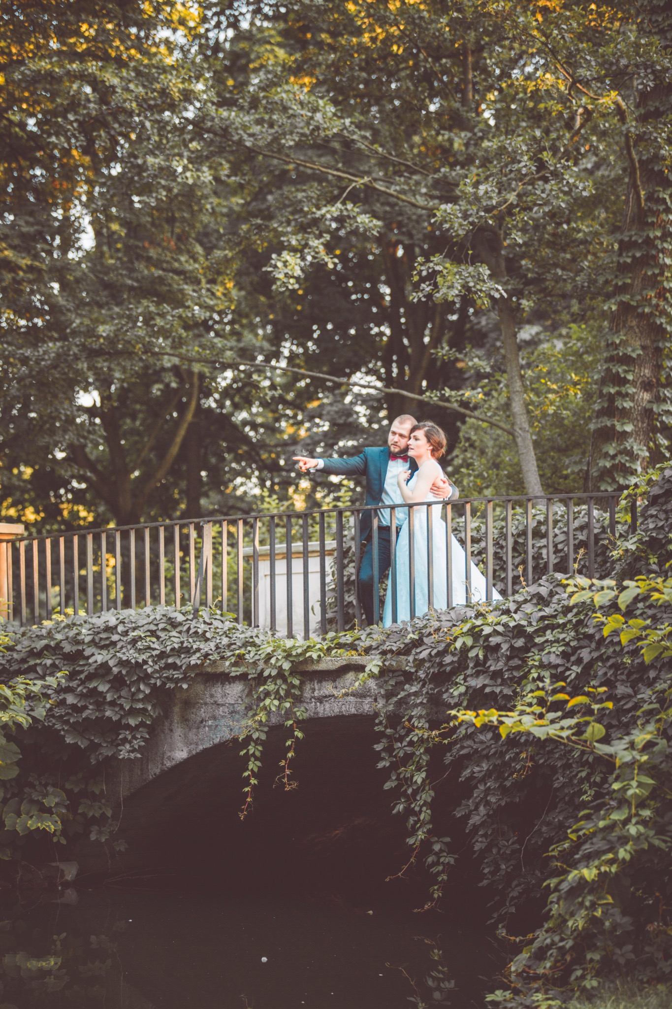 Para młoda w objęciach na kamiennym mostku porośniętym bluszczem, pan młody wskazuje palcem w dal, drzewa w tle.