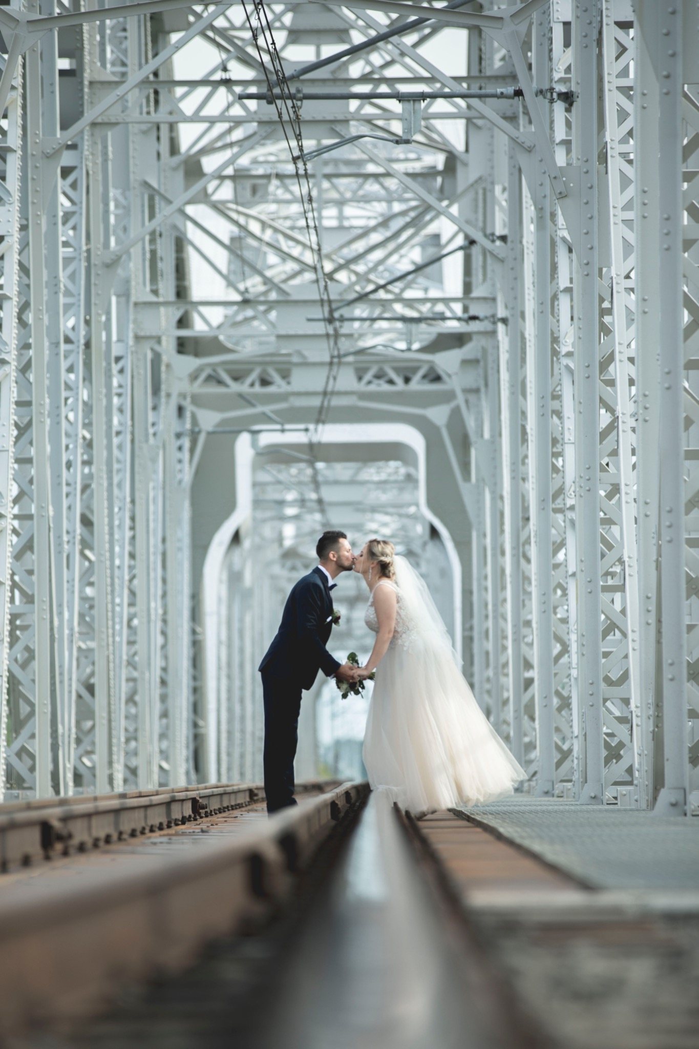 Para młoda całuje się na tle białej, metalowej konstrukcji mostu z widocznymi torami kolejowymi.