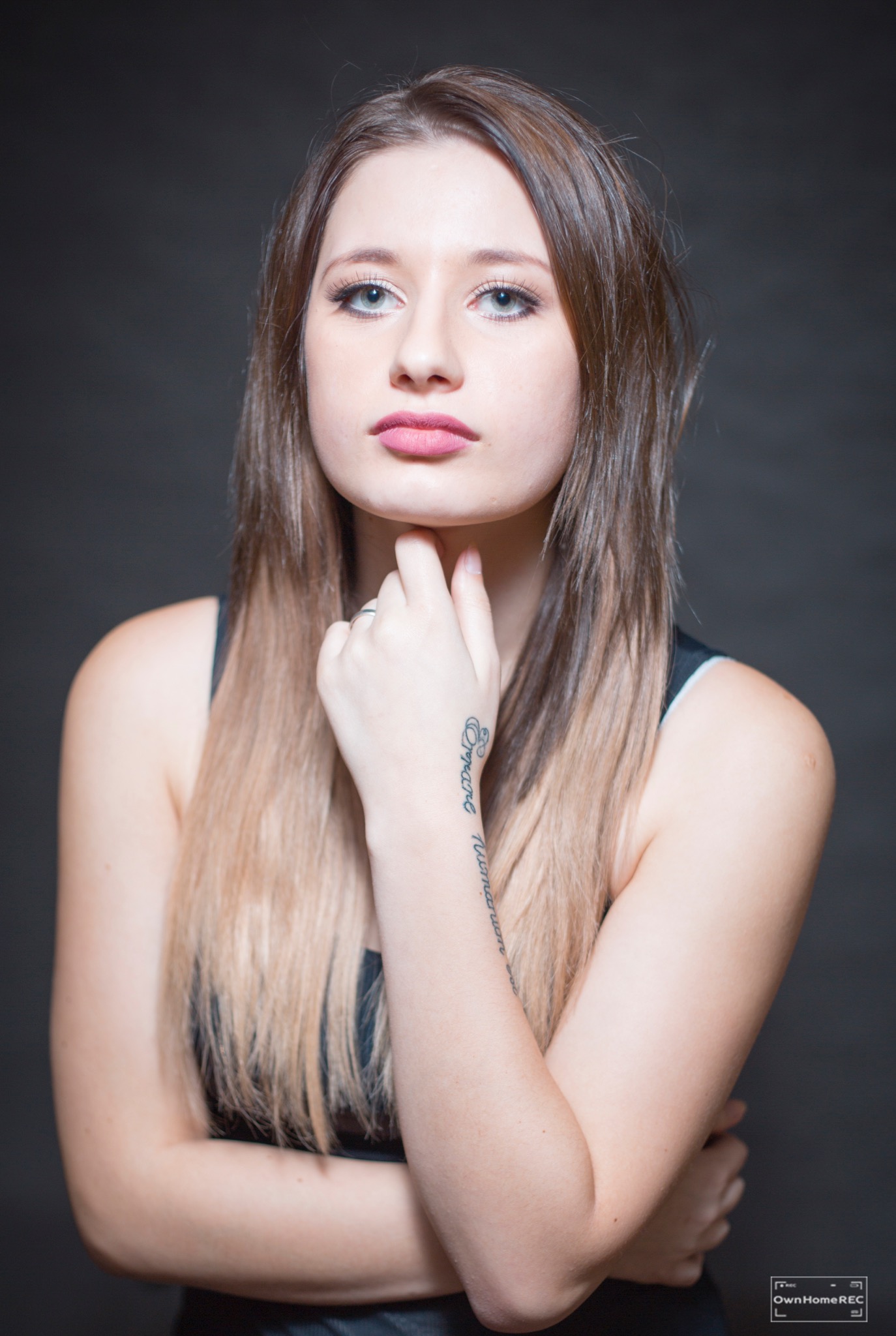 Portret kobiety z ombre włosami, dłoń dotyka szyi, widoczny tatuaż na przedramieniu, neutralne tło.