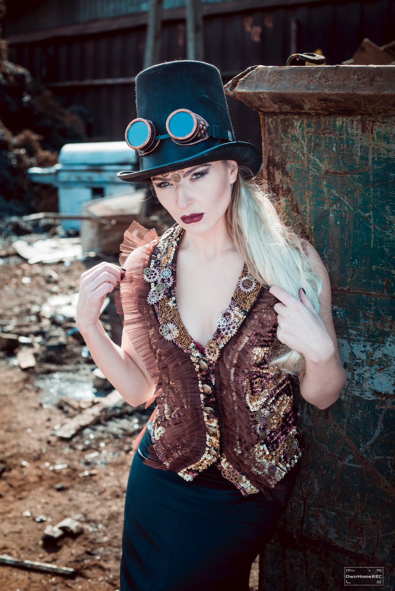 Kobieta w steampunkowym stroju: cylinder z goglami, bogato zdobiona kamizelka z zębatkami, ciemna szminka, pozuje na tle zardzewiałego kontenera i gruzu.