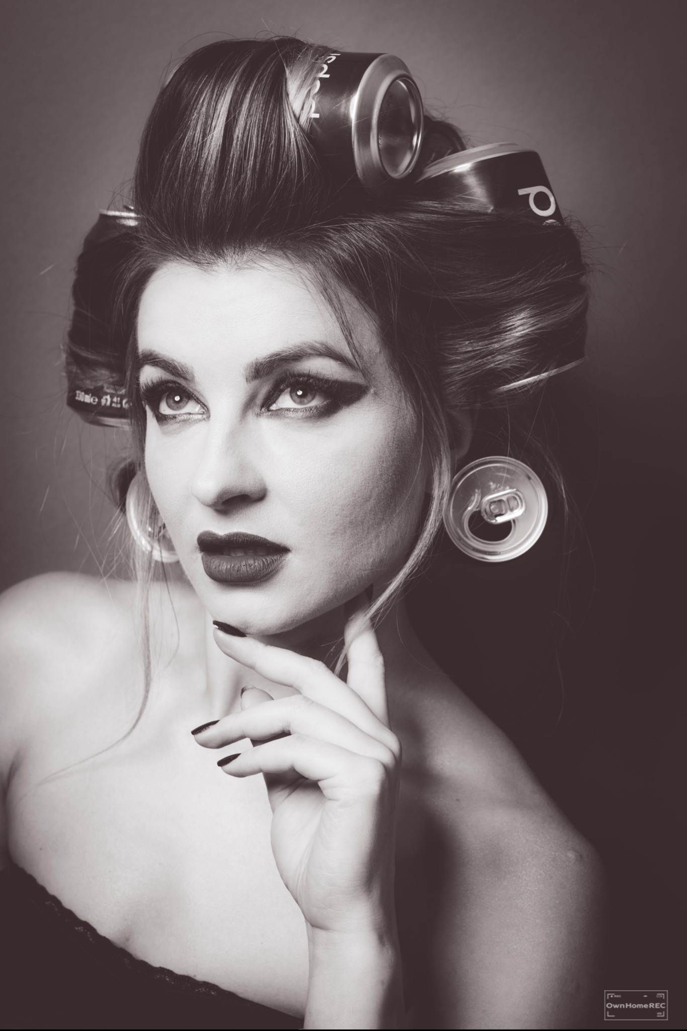 Czarno-biały portret kobiety z pełnym makijażem, włosami upiętymi w puszyste loki z puszek po napojach, które służą jako wałki, i kolczykami z otwartych zawleczek puszek.
