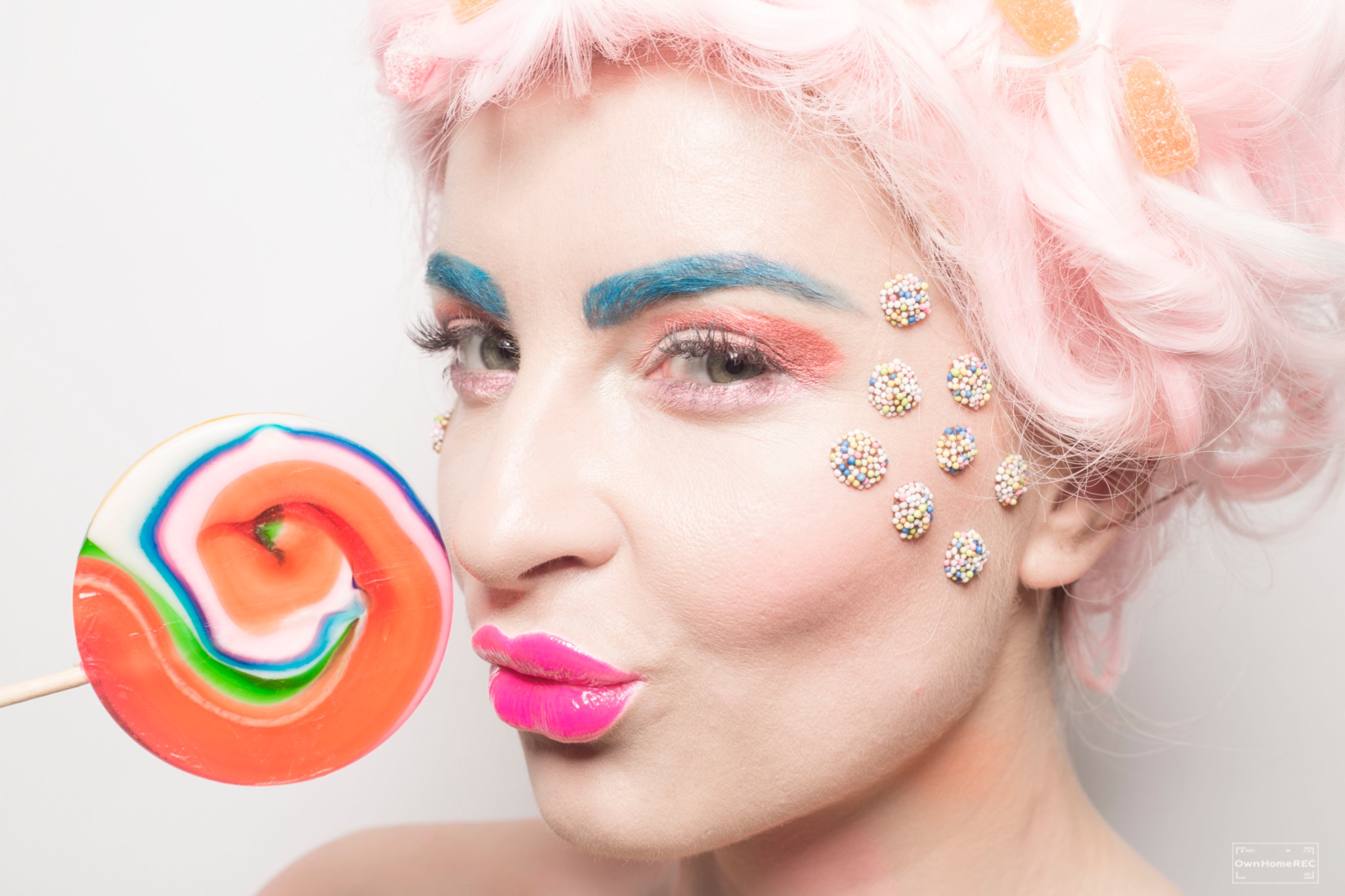 Portret kobiety z różową peruką, niebieskimi brwiami, kolorowym makijażem i lizakiem, ozdobiona posypką cukierniczą na twarzy.