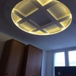 Okrągły sufit podwieszany z podświetleniem LED, tworzący geometryczny wzór krzyża, widoczny w pokoju z dwoma drewnianymi szafami i oknem.