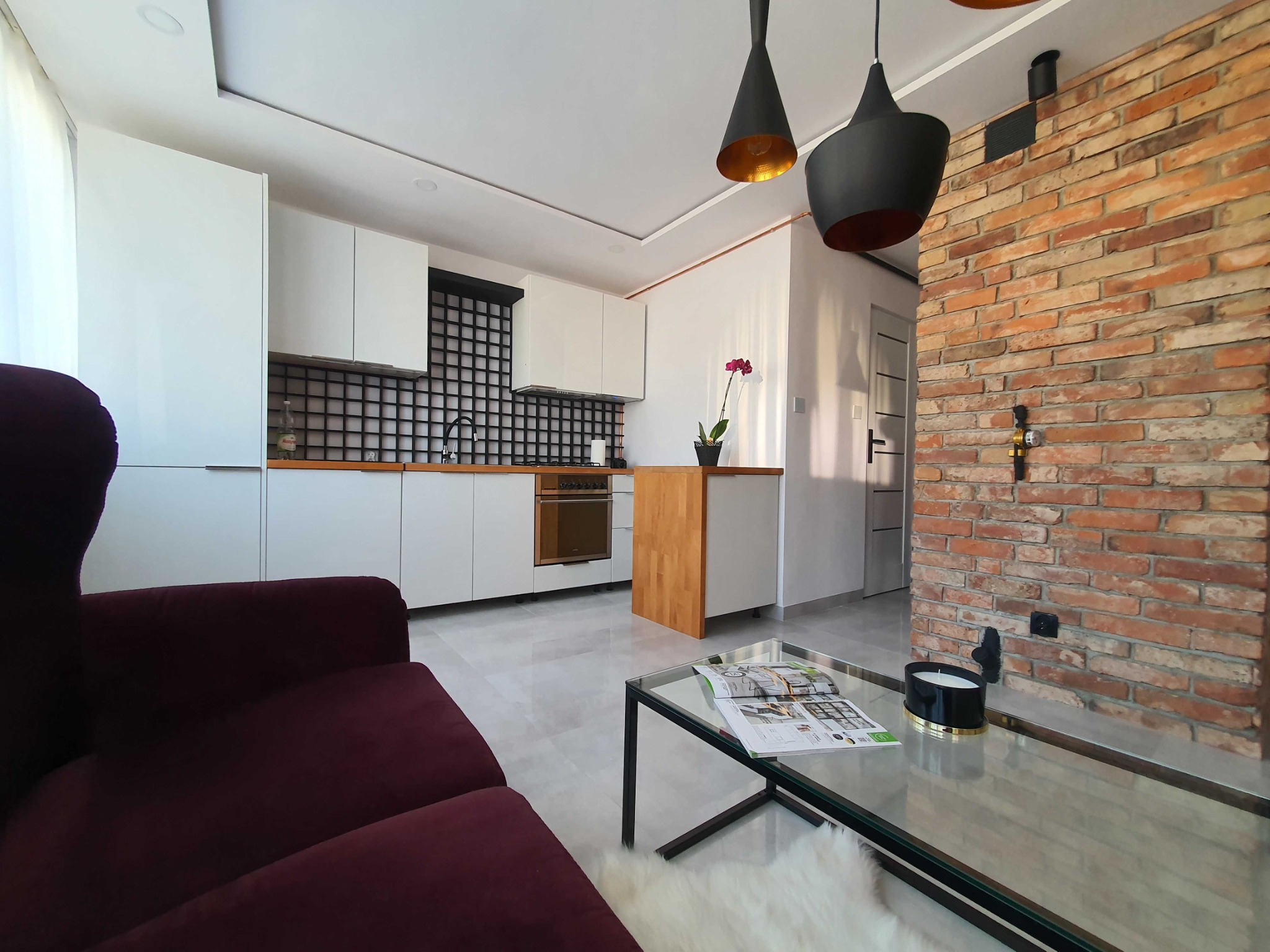 Nowoczesna kuchnia z białymi szafkami, czarną kratką nad blatem, ceglaną ścianą, wiszącymi lampami z złotym wnętrzem i burgundową sofą w salonie