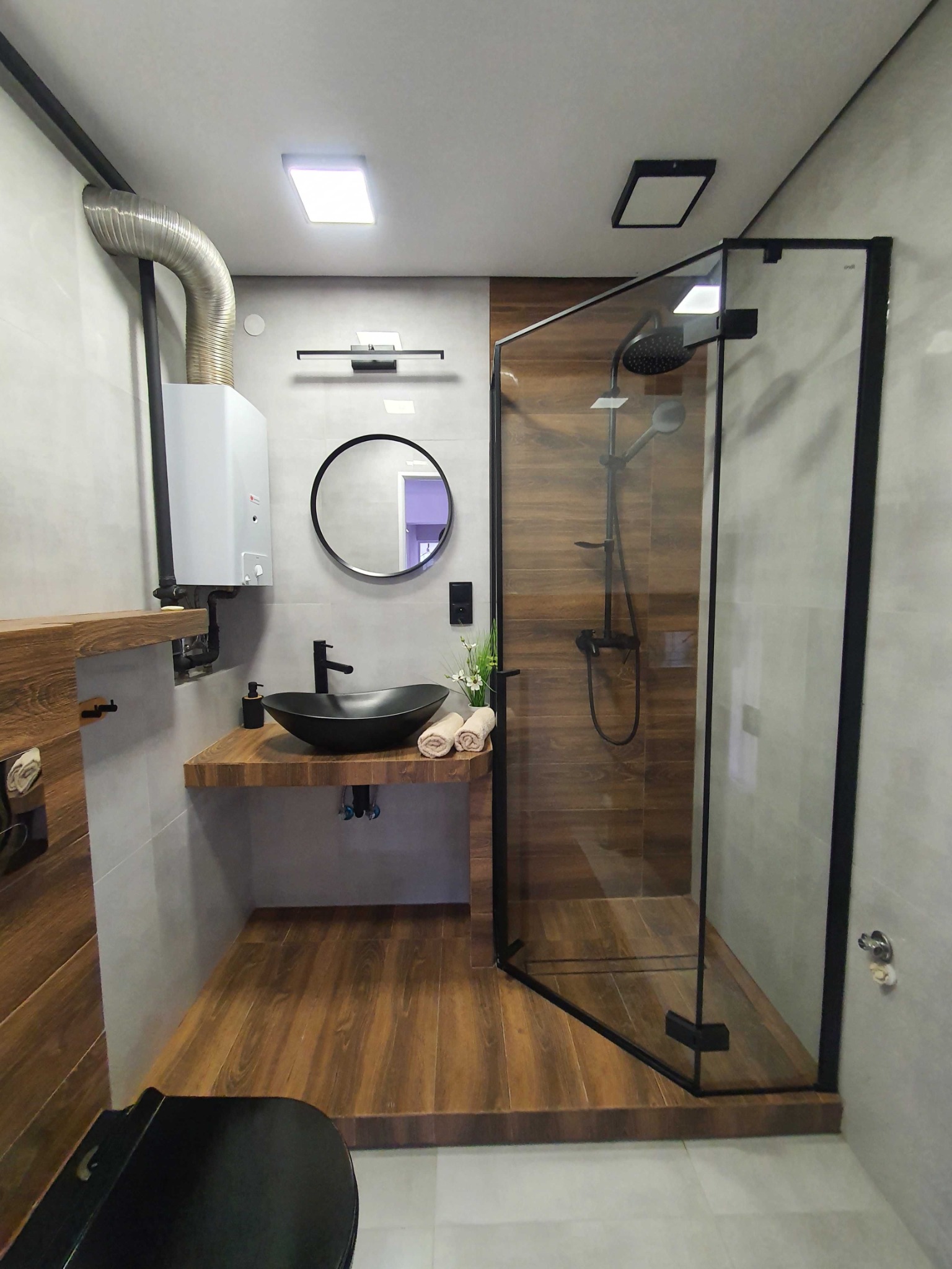 Nowoczesna łazienka z czarną armaturą, kabiną prysznicową typu walk-in z deszczownicą, okrągłym lustrem i umywalką w kształcie misy, drewnopodobne panele ścienne i podłogowe, oraz wiszący piec...