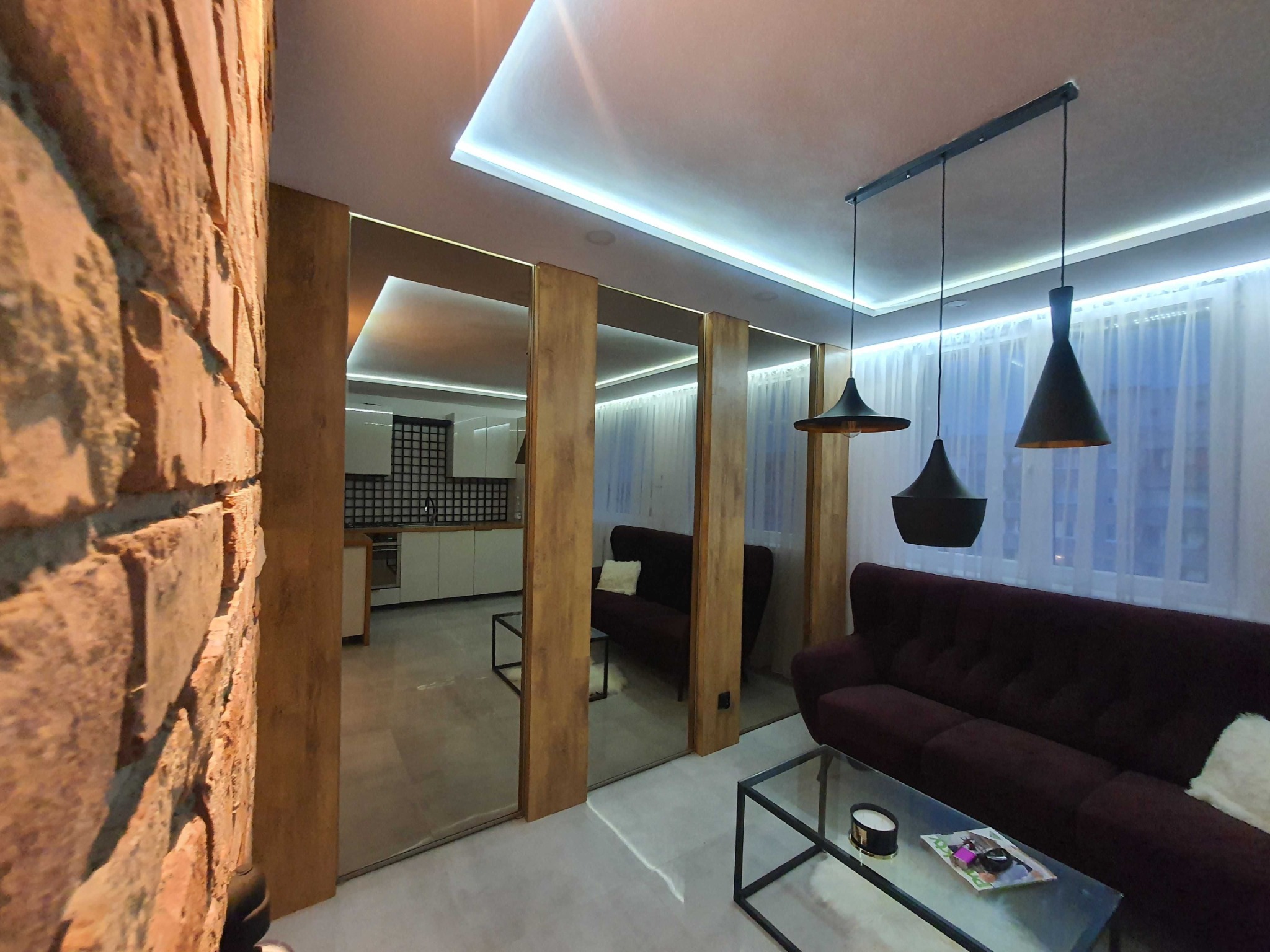 Salon z ceglaną ścianą, lustrzanymi drzwiami przesuwnymi, burgundową sofą i nowoczesnym oświetleniem LED podsufitowym.