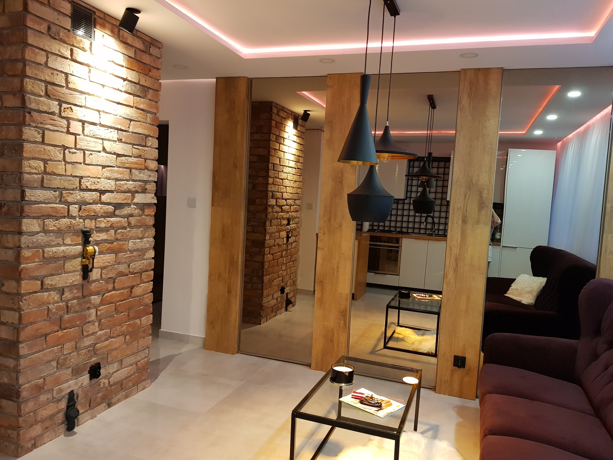 Nowoczesne wnętrze: ceglana ściana z oświetleniem punktowym, lustrzane panele z drewnianymi wstawkami, wiszące czarne lampy, fioletowa sofa i szklane stoliki kawowe.