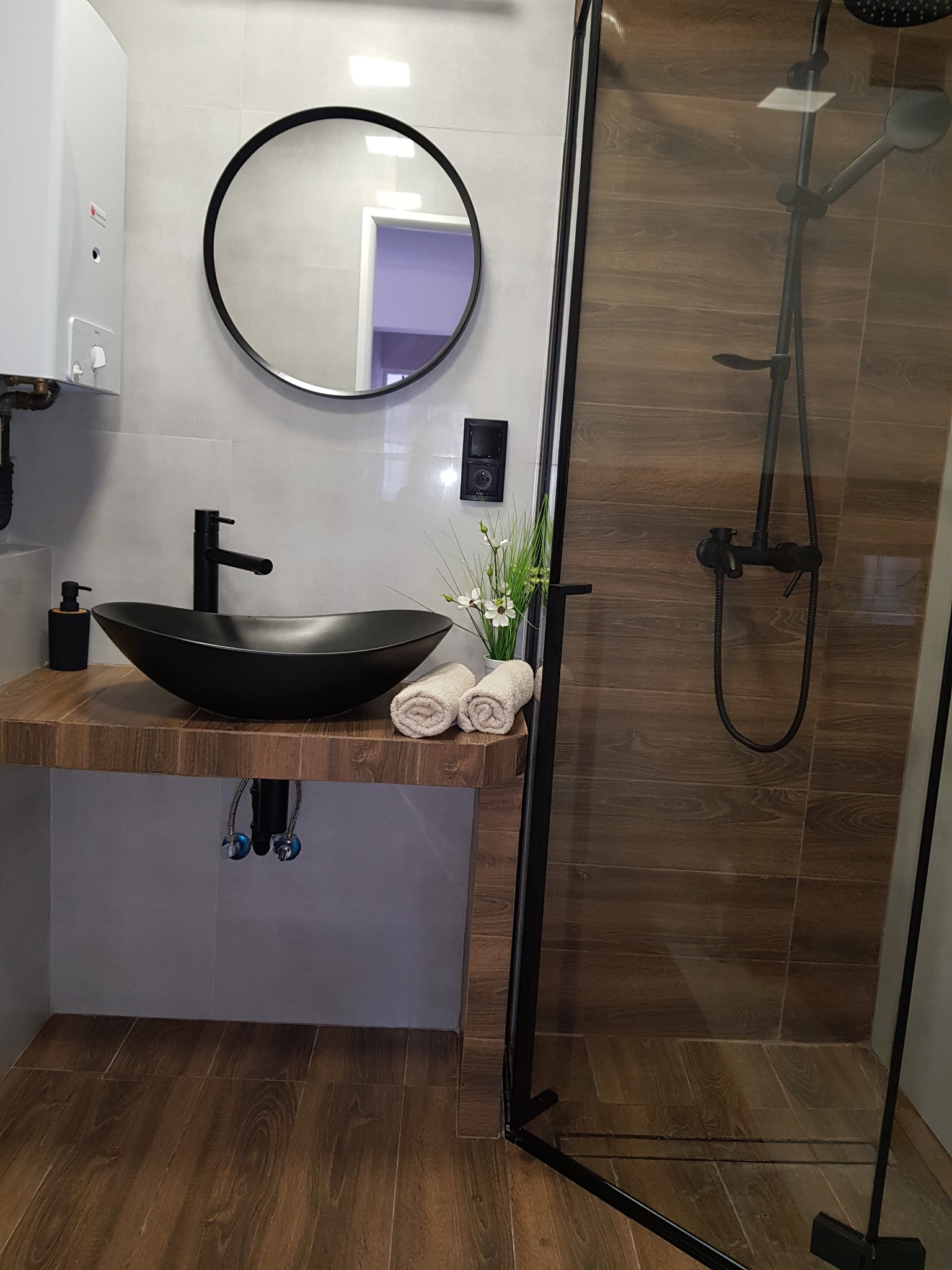 Nowoczesna łazienka z czarną, owalną umywalką nablatową, drewnianym blatem, okrągłym lustrem i kabiną prysznicową typu walk-in z brązowymi płytkami imitującymi drewno.