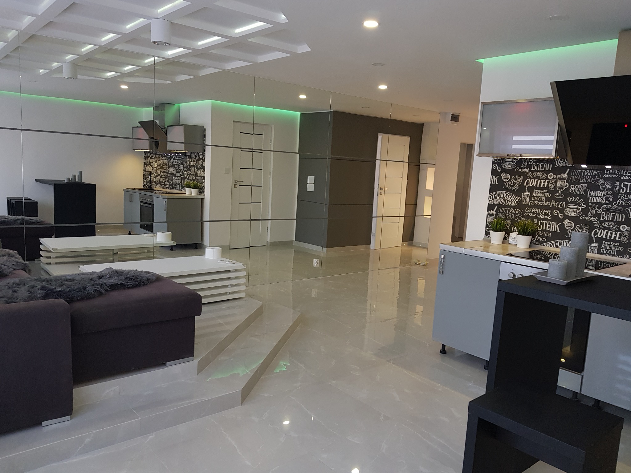 Przestronne wnętrze mieszkania z lustrzaną ścianą, widok na kuchnię z czarno-białą tapetą, szare meble i subtelne oświetlenie LED