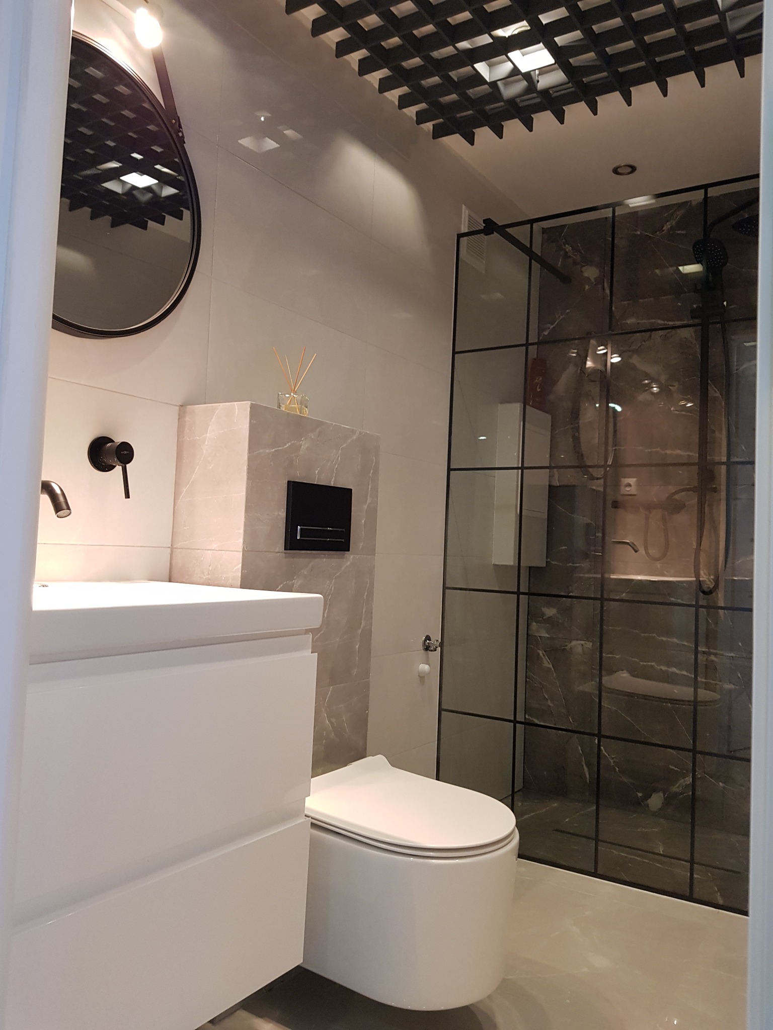 Nowoczesna łazienka z kabiną prysznicową w stylu loft, umywalką z szafką w kolorze białym, okrągłym lustrem i czarną armaturą na tle płytek imitujących marmur.