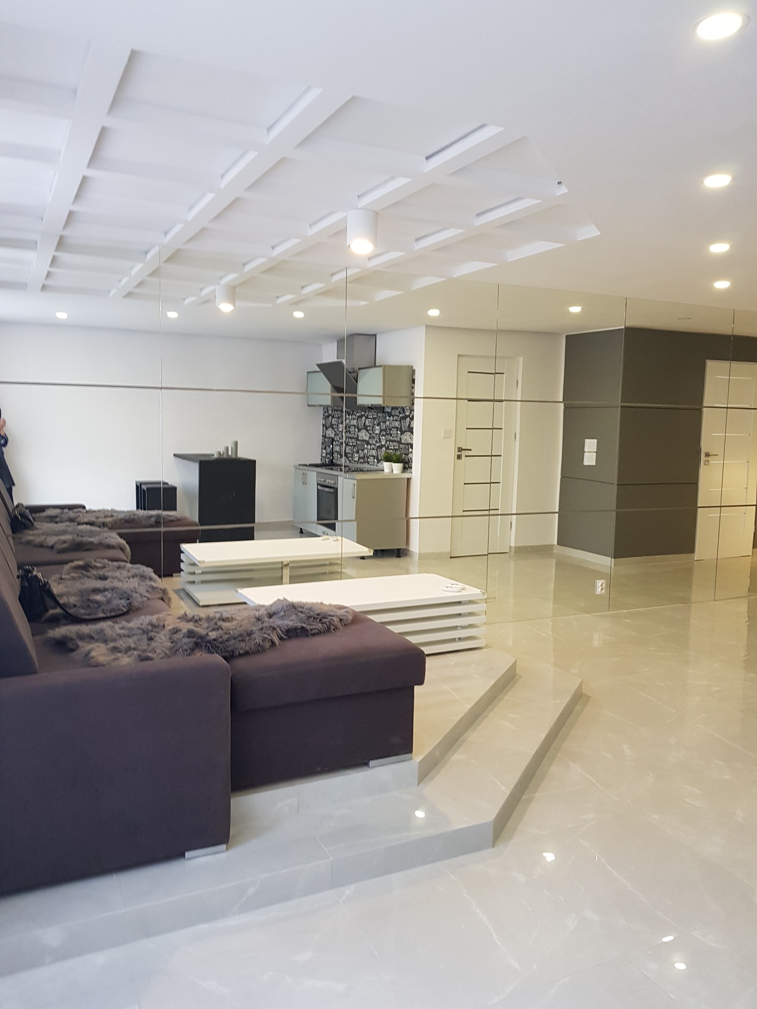 Nowoczesny salon z lustrzanymi ścianami, brązową sofą z futrzanymi poduszkami, białym stolikiem kawowym i schodami, widok na kuchnię z czarno-białą mozaiką nad blatem.