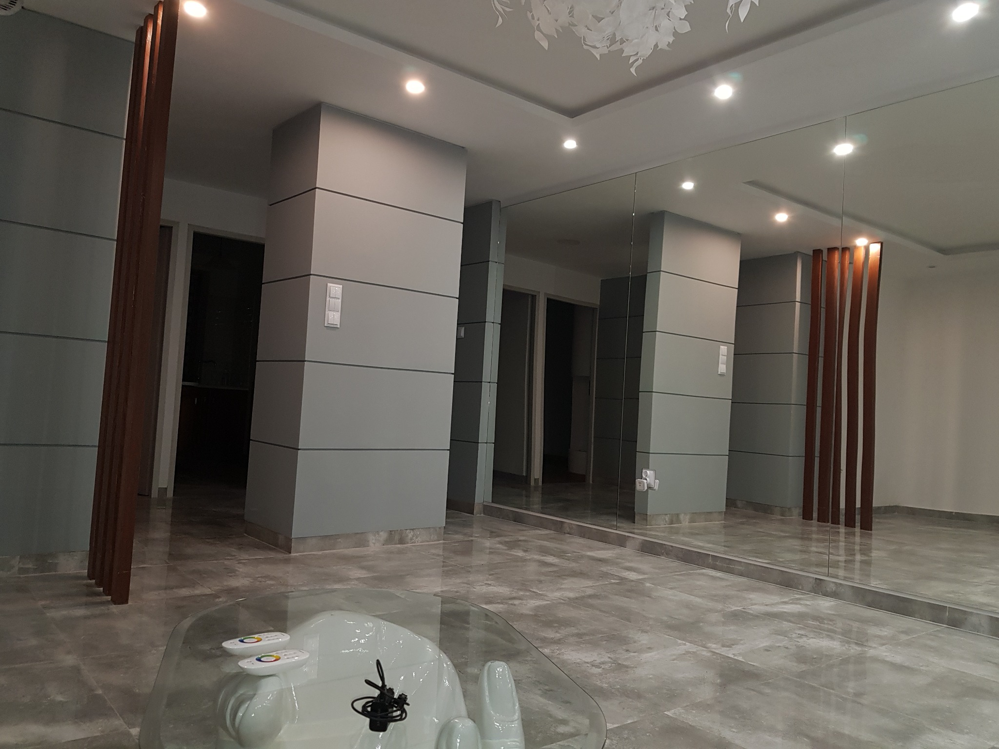 Nowoczesne wnętrze: szare ściany z poziomymi panelami, lustro na całej ścianie odbijające drewniane elementy dekoracyjne, szklany stolik kawowy z pilotami i mikrofonem, podłoga z płytek imitujących...