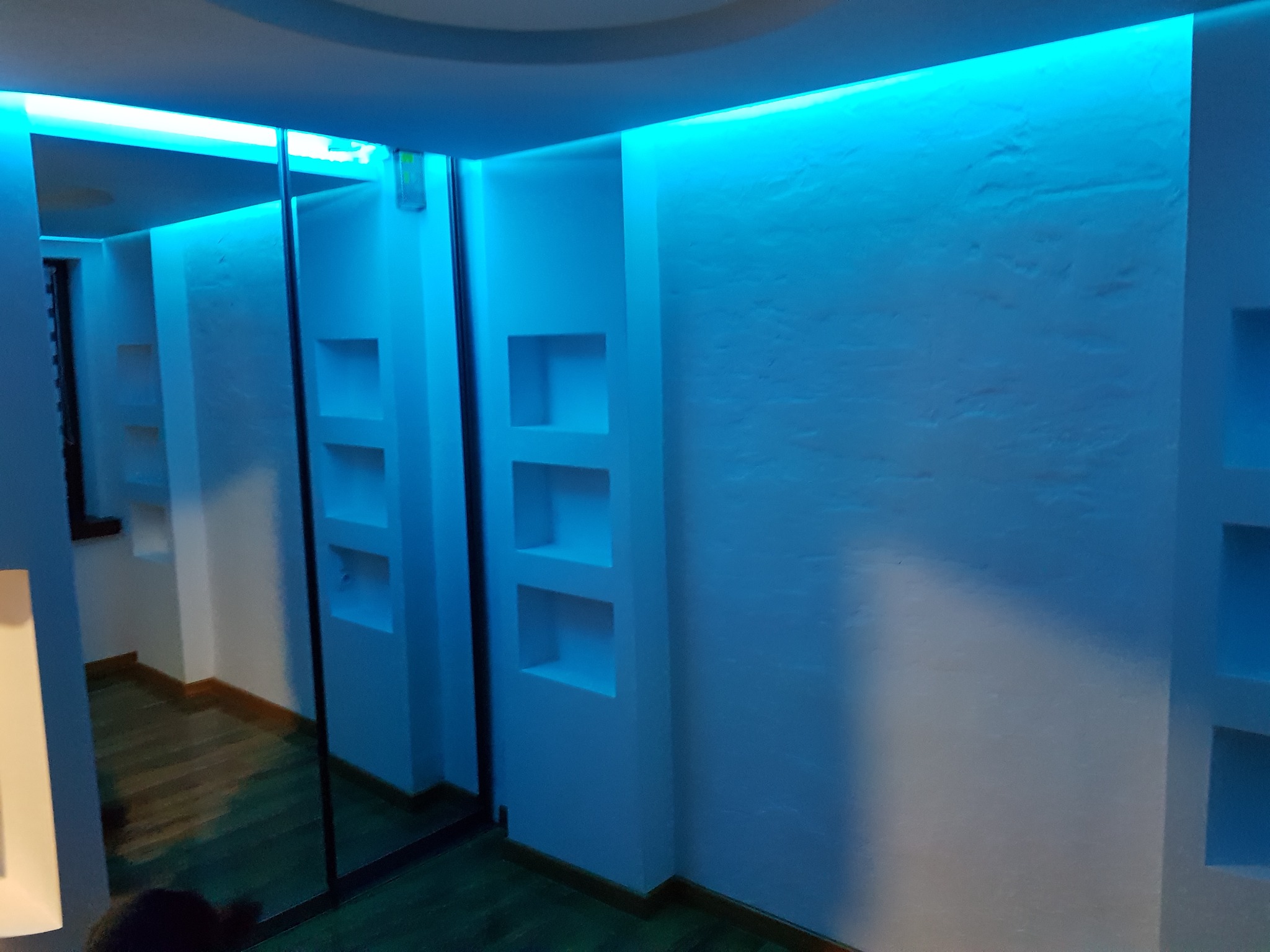 Nowoczesne wykończenie wnętrza: biała ściana z wnękami oświetlona niebieskim światłem LED, przesuwne drzwi z lustrem odbijające wnętrze pokoju.