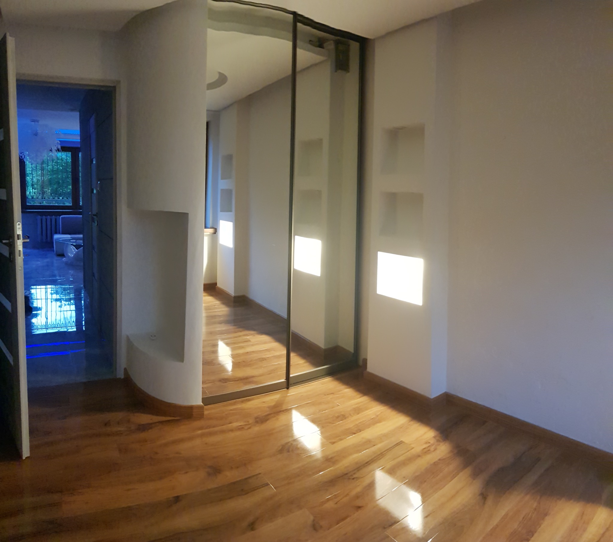 Jasne wnętrze z drewnianą podłogą i wnękami w ścianach, widoczne przesuwne drzwi do kolejnego pomieszczenia z niebieskim oświetleniem.