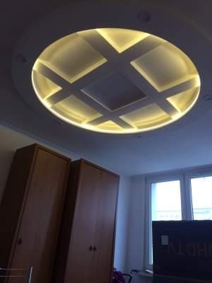Okrągły sufit podwieszany z podświetleniem LED, tworzący geometryczny wzór krzyża, widoczny w pokoju z dwoma drewnianymi szafami i oknem.