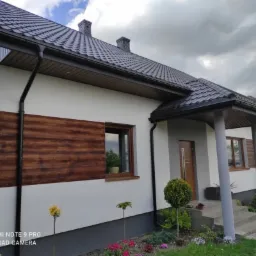 Jasny dom jednorodzinny z ciemną dachówką i drewnianą okładziną elewacyjną, widok z ogrodu z ozdobnymi krzewami i kwiatami.