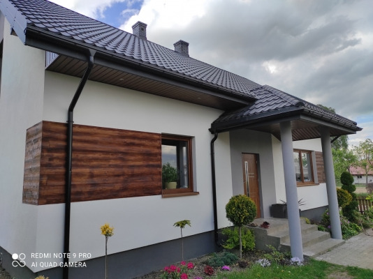 Jasny dom jednorodzinny z ciemną dachówką i drewnianą okładziną elewacyjną, widok z ogrodu z ozdobnymi krzewami i kwiatami.