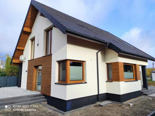 Dwukondygnacyjny dom z jasną elewacją, drewnianymi akcentami i ciemnym dachem, uchwycony w słoneczny dzień z niskiej perspektywy.