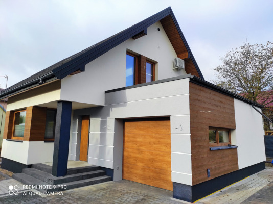 Nowoczesny dom jednorodzinny z elewacją w odcieniach szarości i brązu, z garażem i klimatyzatorem na ścianie, widok z poziomu chodnika z kostki brukowej.