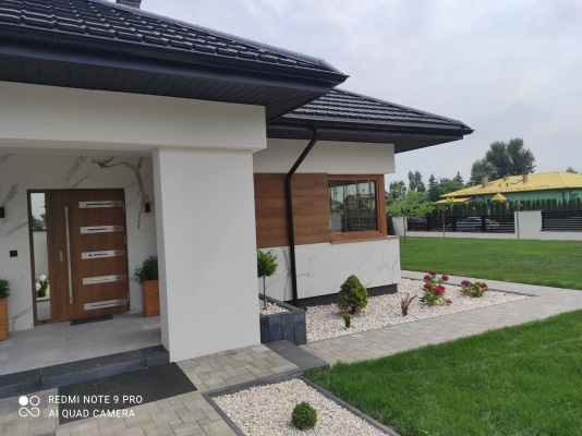 Nowoczesny dom jednorodzinny z elewacją w odcieniach bieli i drewna, widok z zewnątrz, z kostką brukową i elementami zieleni w ogrodzie.