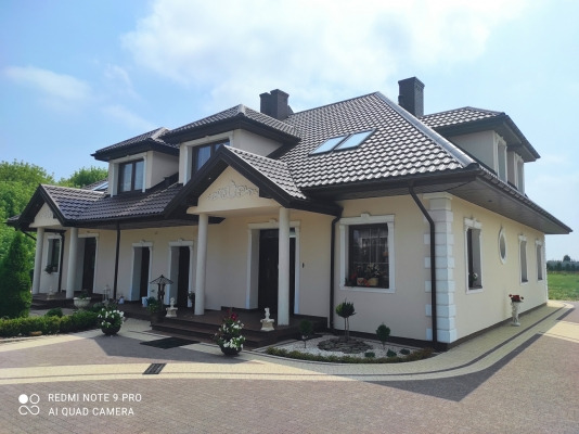 Dwukondygnacyjny dom jednorodzinny z beżową elewacją, brązowym dachem i kolumnami przy wejściu, otoczony zadbanym ogrodem i brukowanym podjazdem w słoneczny dzień.