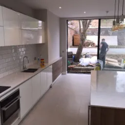 Nowoczesna, jasna kuchnia z białymi szafkami i szarymi płytkami na ścianie, widok na ogród przez duże okno, w którym stoi mężczyzna