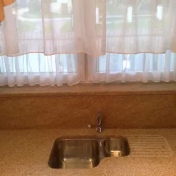 Blat kuchenny z konglomeratu z dwukomorowym zlewem ze stali nierdzewnej i ociekaczem, zamontowany pod oknem z firankami.