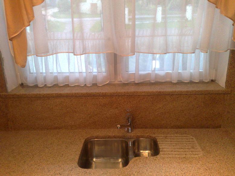 Blat kuchenny z konglomeratu z dwukomorowym zlewem ze stali nierdzewnej i ociekaczem, zamontowany pod oknem z firankami.